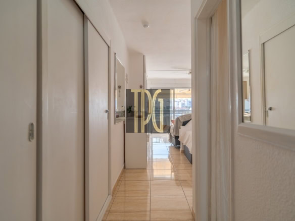 Bright hotel hallway leading to a sunlit bedroom with a bed at the far end, white walls, and glossy tiled floor. Watermark in center reads 'DPG'.