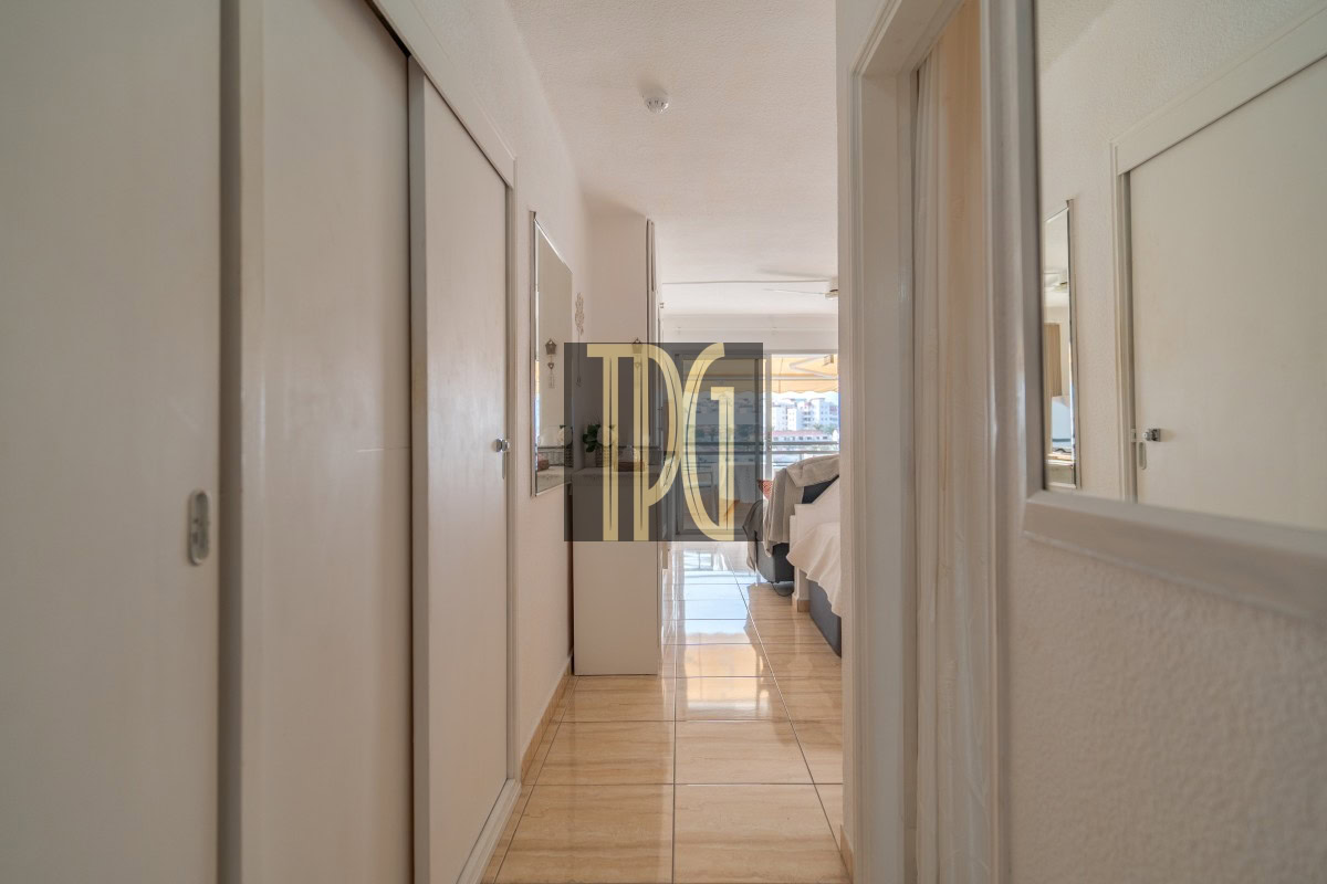 Bright hotel hallway leading to a sunlit bedroom with a bed at the far end, white walls, and glossy tiled floor. Watermark in center reads 'DPG'.