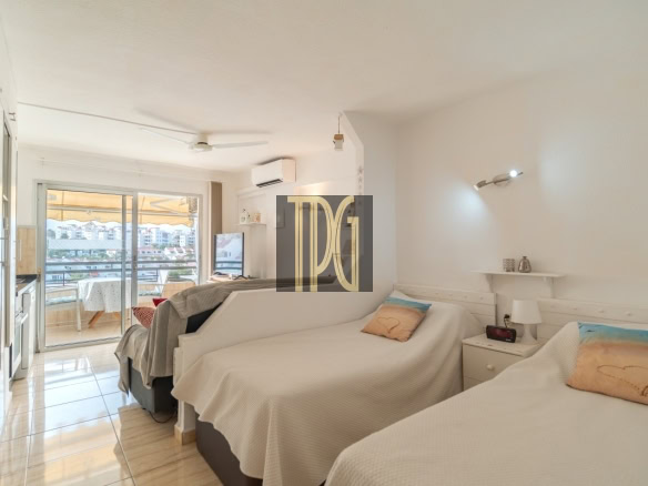 Bright hotel room with two single beds, white bedding, and a small nightstand; a balcony opens to a city view in the background.