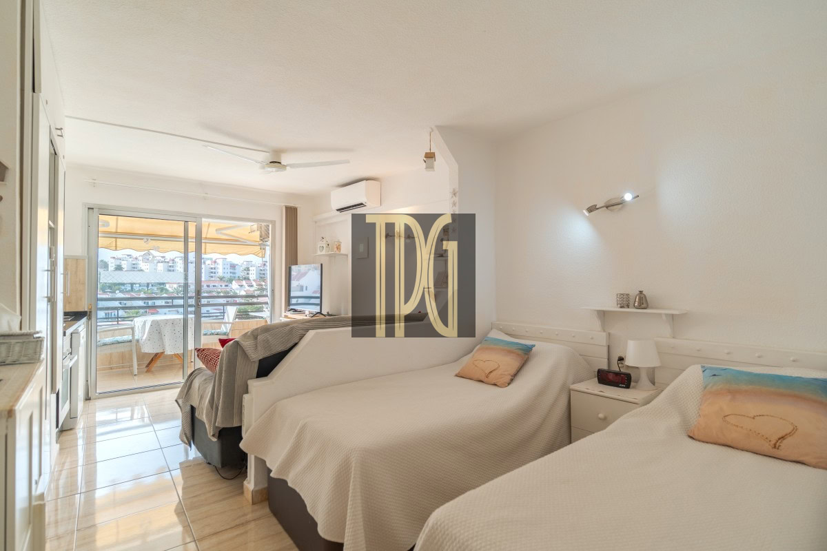 Bright hotel room with two single beds, white bedding, and a small nightstand; a balcony opens to a city view in the background.