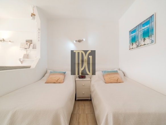 Twin single beds with white bedding and pastel beach pillows, separated by a small nightstand in a bright, minimalist hotel room.