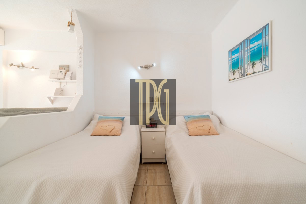 Twin single beds with white bedding and pastel beach pillows, separated by a small nightstand in a bright, minimalist hotel room.