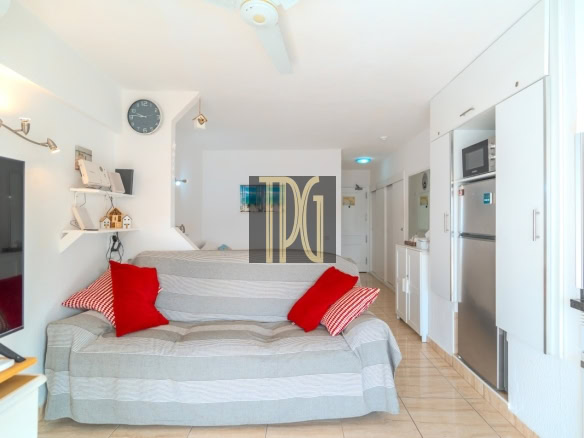 Bright open-plan living space with a gray striped sofa and red cushions, wall clock, TV on a stand, and a compact kitchen with stainless appliances to the right.