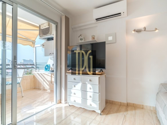 Sunlit living room with a flat-screen TV on a white dresser and sliding glass doors to a balcony.