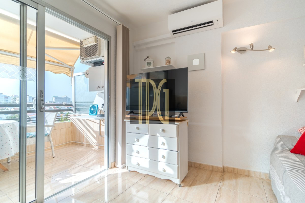 Sunlit living room with a flat-screen TV on a white dresser and sliding glass doors to a balcony.