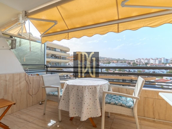 Balcony seating area with a round table under a yellow awning and two wicker chairs overlooking a cityscape.