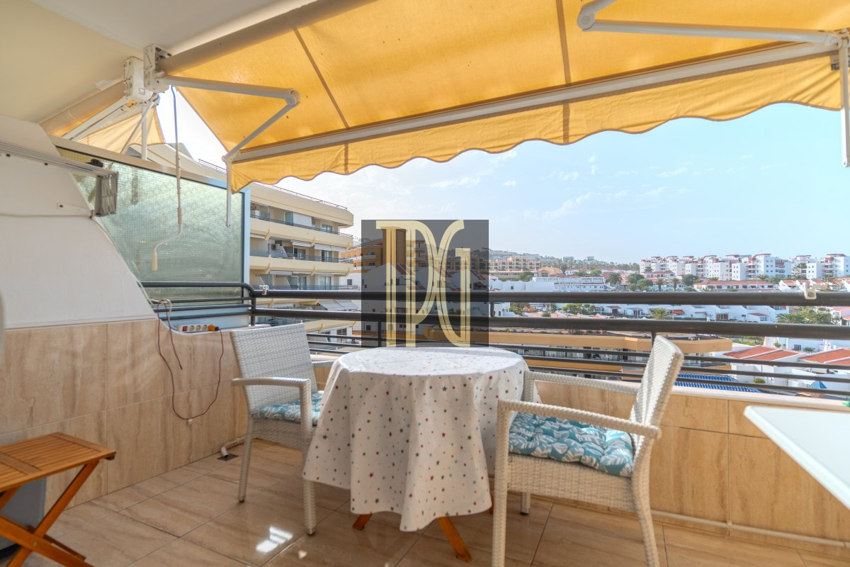 Balcony seating area with a round table under a yellow awning and two wicker chairs overlooking a cityscape.
