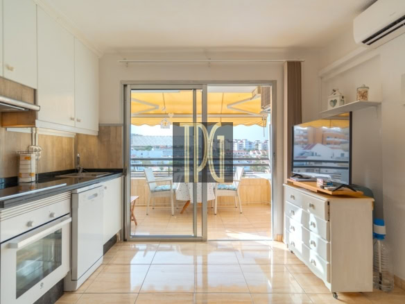Bright kitchen with white cabinets leading to a sunny balcony overlooking a cityscape; dishwasher and oven visible.