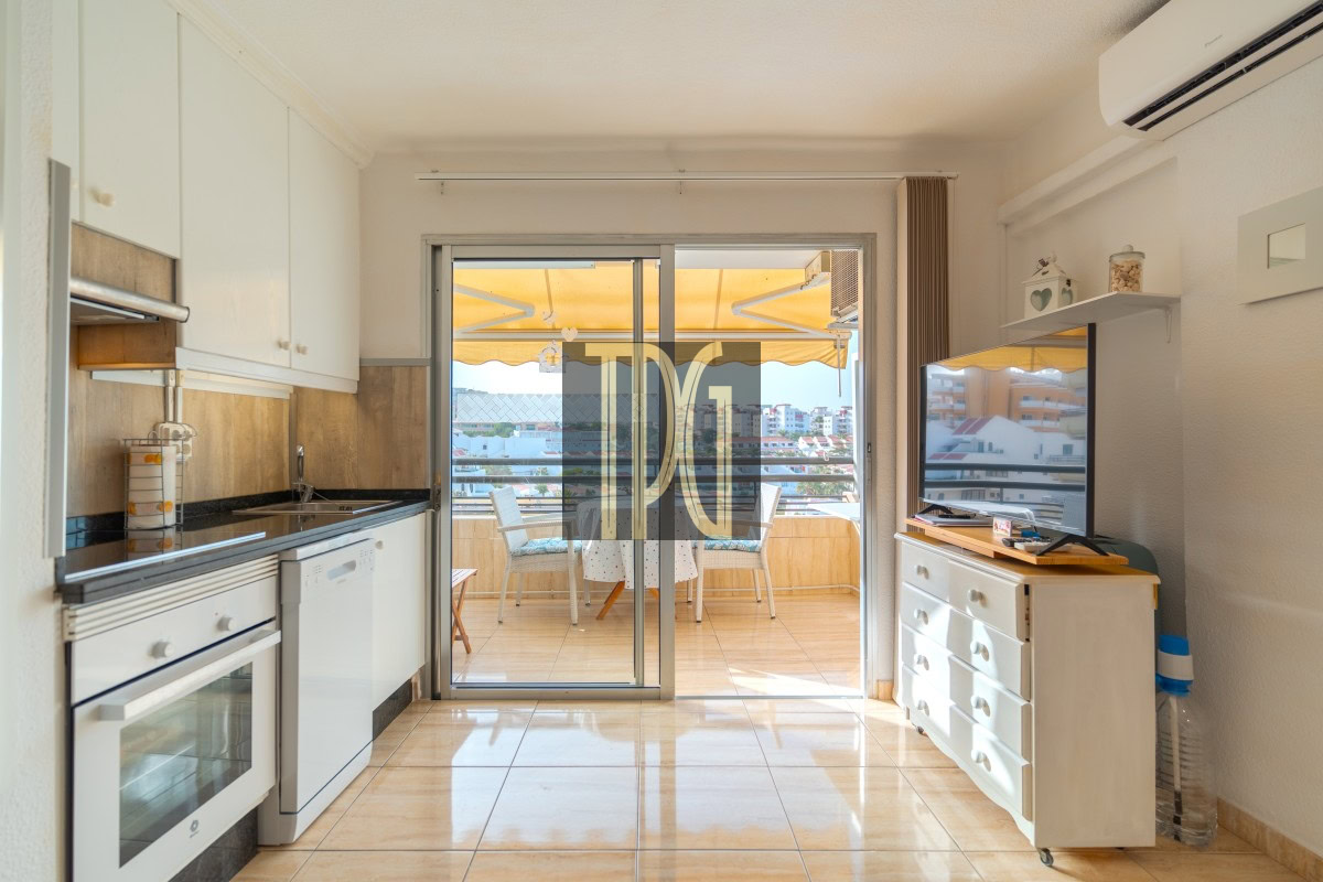 Bright kitchen with white cabinets leading to a sunny balcony overlooking a cityscape; dishwasher and oven visible.