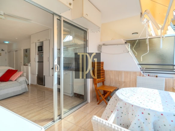 Balcony with round table covered by a star-patterned cloth, wicker chairs, and a folding wooden chair; sliding glass doors lead to a bright interior.