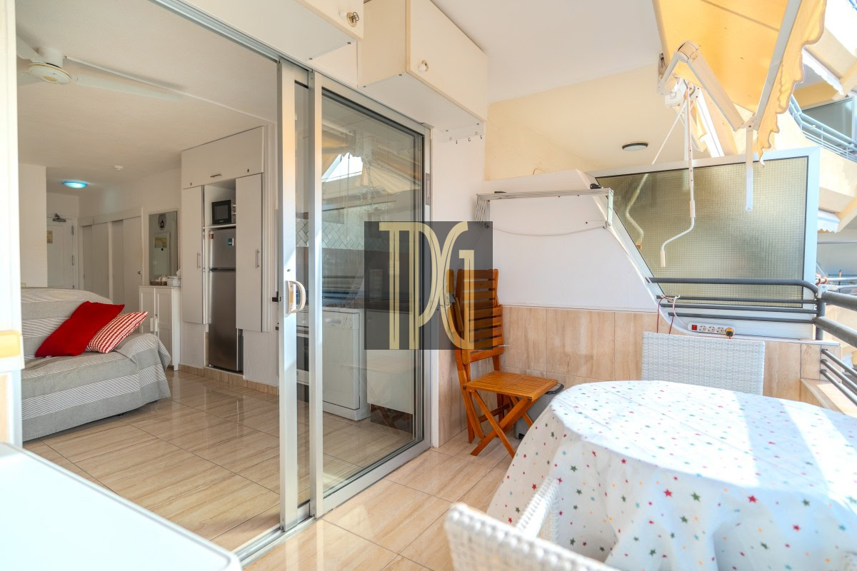 Balcony with round table covered by a star-patterned cloth, wicker chairs, and a folding wooden chair; sliding glass doors lead to a bright interior.