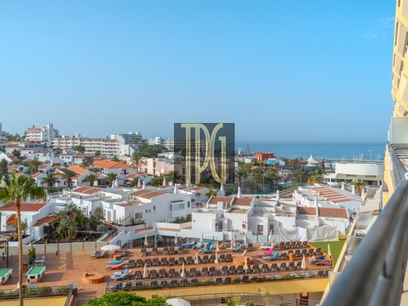 Coastal resort scene with white Mediterranean-style buildings, palm trees, and a pool terrace with sun loungers facing the sea.