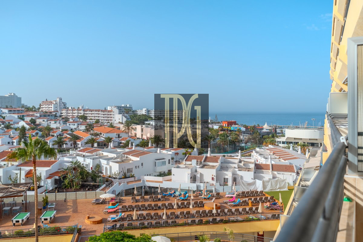 Coastal resort scene with white Mediterranean-style buildings, palm trees, and a pool terrace with sun loungers facing the sea.