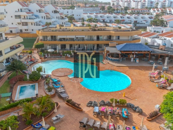 Resort pool area with two blue pools, sun loungers, and yellow hotel buildings surrounding a tiled terrace.
