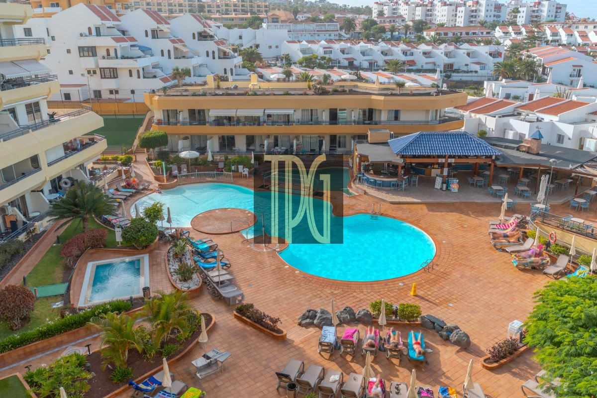 Resort pool area with two blue pools, sun loungers, and yellow hotel buildings surrounding a tiled terrace.