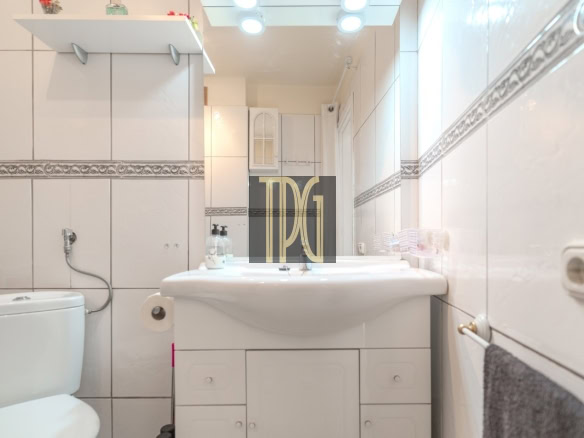 Bright white-tiled bathroom with a wide sink, toilet to the left, and a mirrored vanity cabinet, illuminated by ceiling lights.