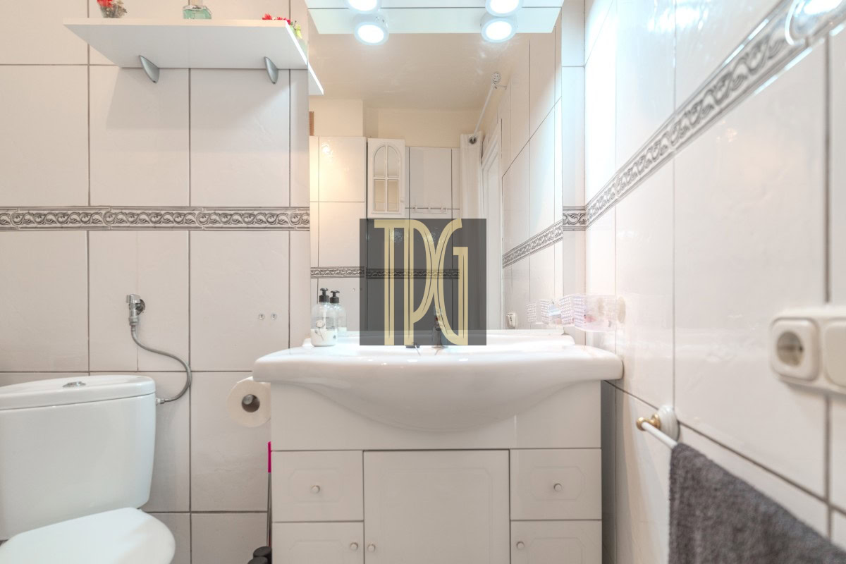 Bright white-tiled bathroom with a wide sink, toilet to the left, and a mirrored vanity cabinet, illuminated by ceiling lights.