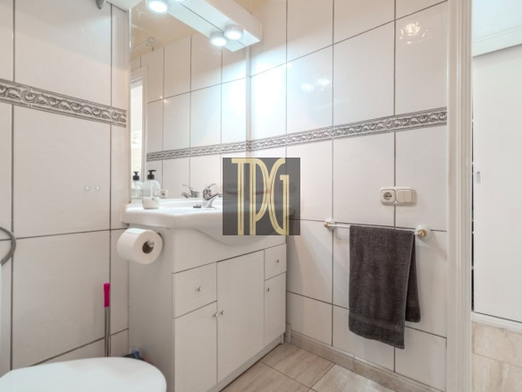 White-tiled bathroom with a white sink and mirrored cabinet, soap dispensers, and a gray towel on a wall bar