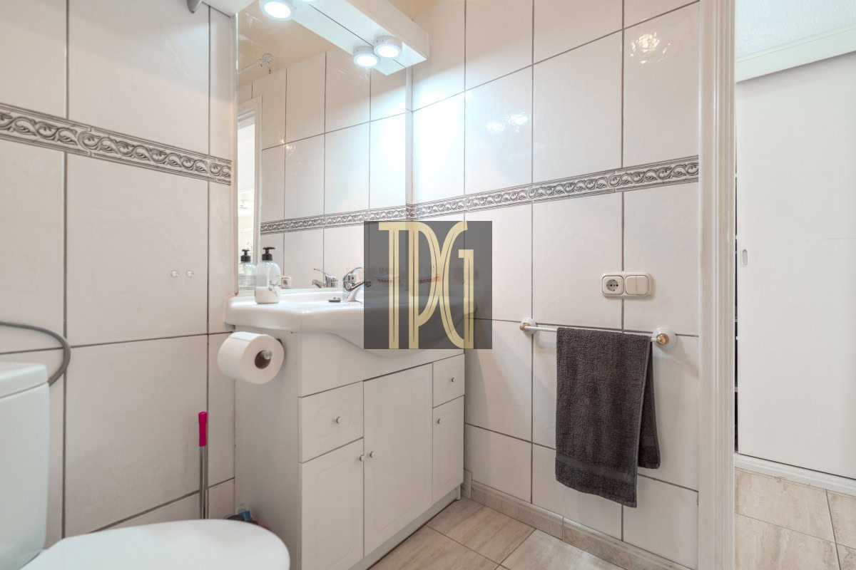 White-tiled bathroom with a white sink and mirrored cabinet, soap dispensers, and a gray towel on a wall bar