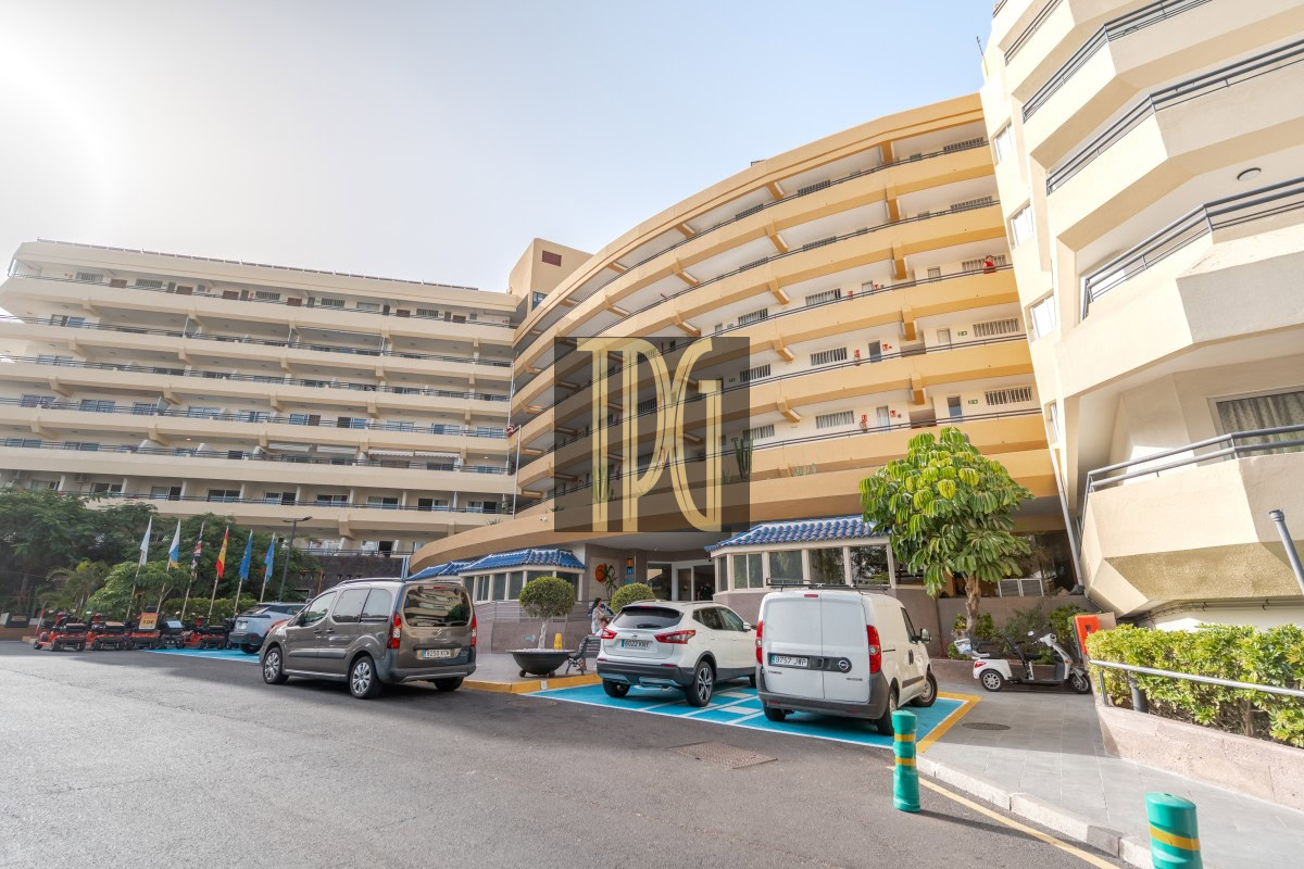 Exterior view of a large multi-story building with curved wings and a front entrance, a row of parked cars, and a tree near the sidewalk; flags line the left side. The scene is bright and sunny, with a blue-tiled entrance canopy visible under the curved structure.