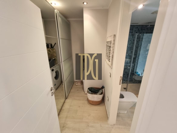 Hallway in a modern apartment showing a front-loading washing machine in a glass-front cabinet, with a laundry basket on the floor and a doorway to a tiled bathroom with a glass shower door.