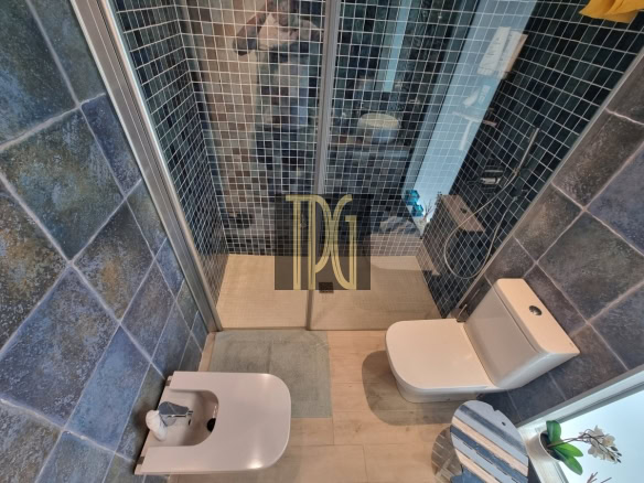 Modern bathroom: glass shower with blue mosaic tiles, and white toilet and bidet with a small striped table and plant nearby.