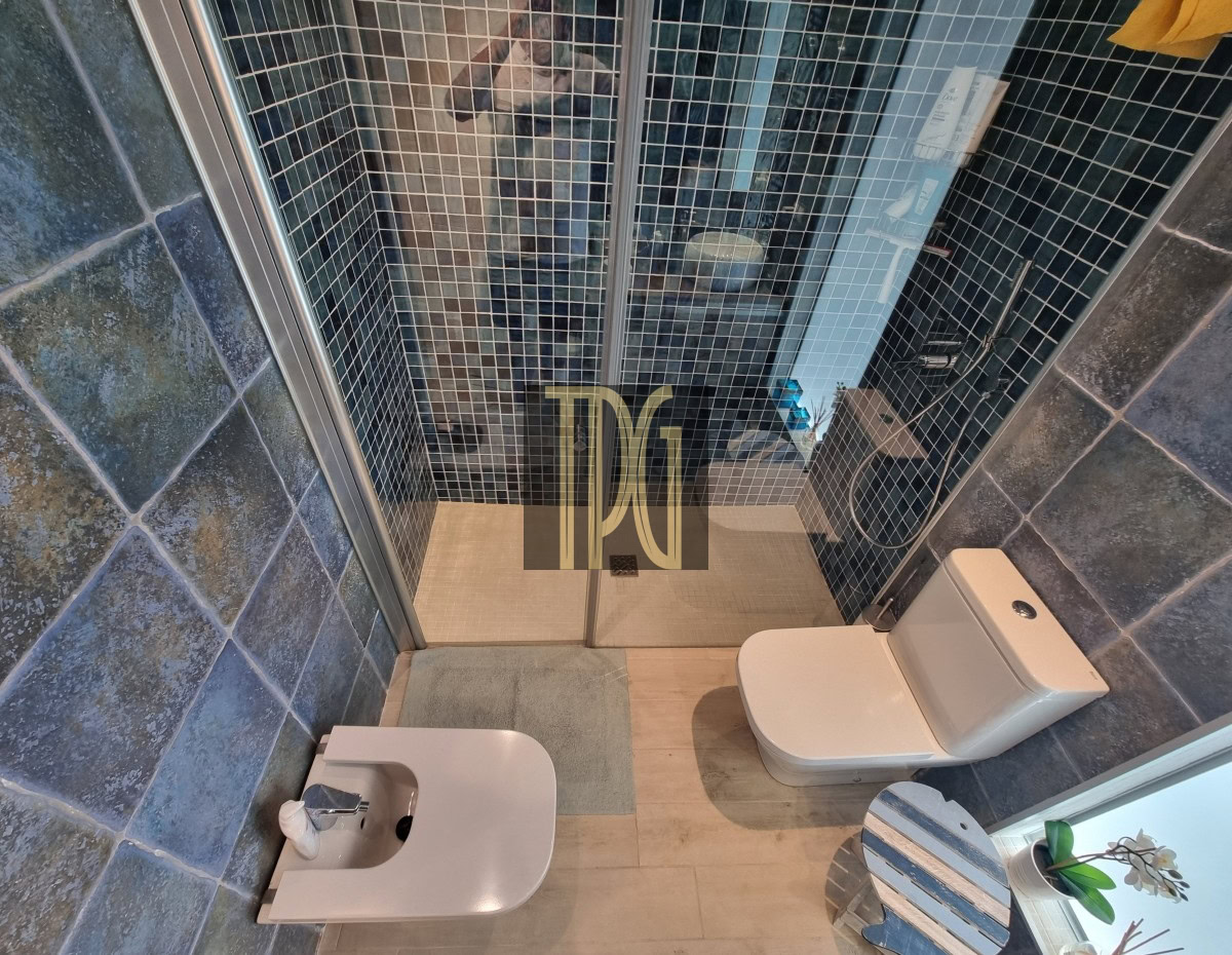 Modern bathroom: glass shower with blue mosaic tiles, and white toilet and bidet with a small striped table and plant nearby.