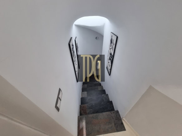 Narrow stone staircase descends between white walls, with black-framed art on either side and a bright arched opening ahead.