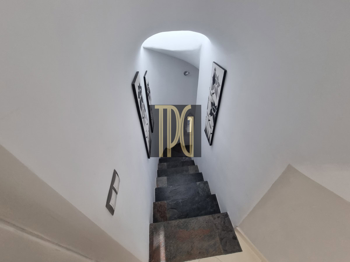Narrow stone staircase descends between white walls, with black-framed art on either side and a bright arched opening ahead.