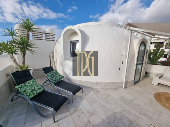 Rooftop terrace with two black lounge chairs and tropical cushions beside a white curved building, under a blue sky.