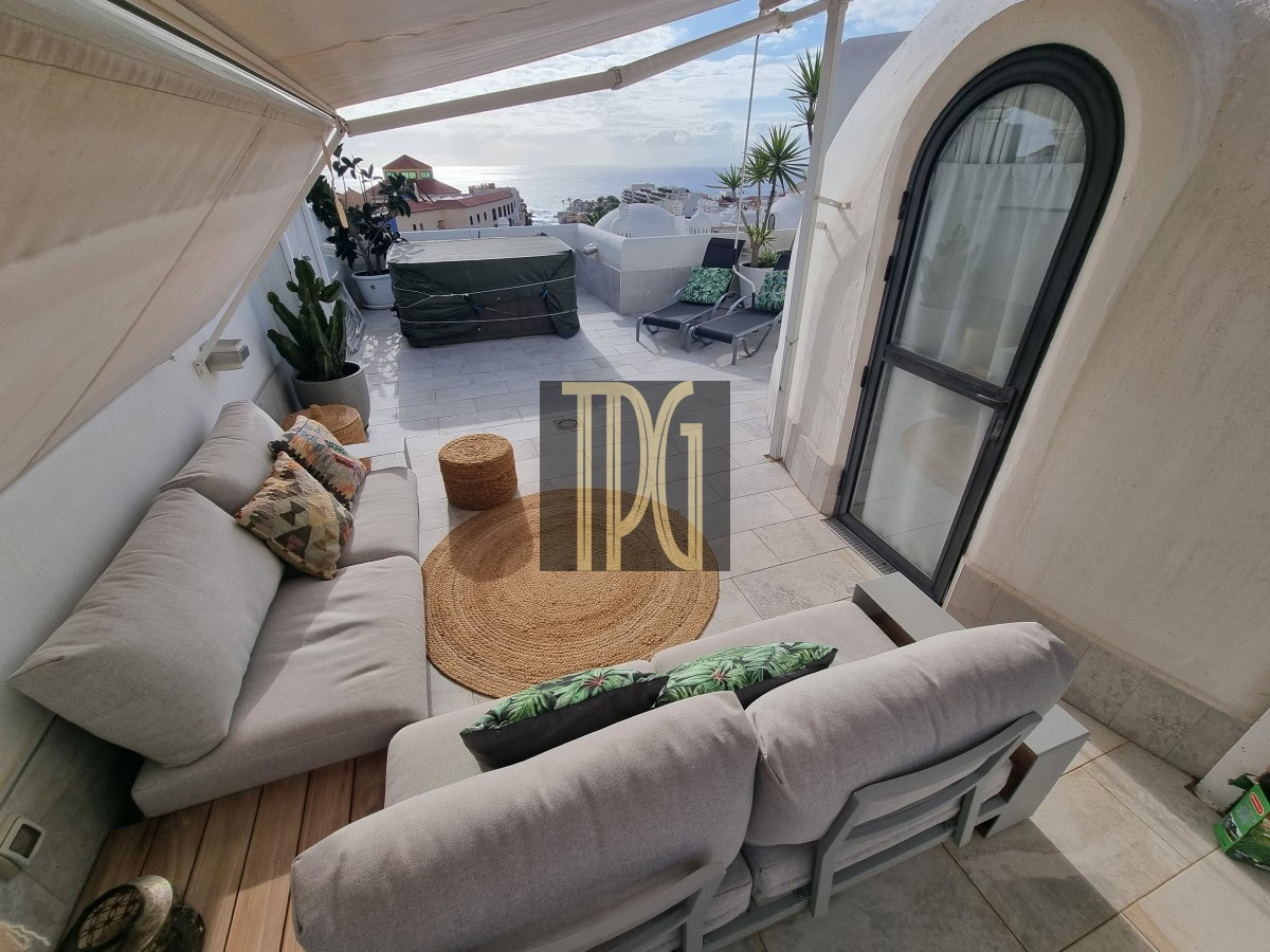 Rooftop terrace with gray cushioned sectional, round jute rug, wicker pouf, and potted plants; sun loungers and sea view in the distance.