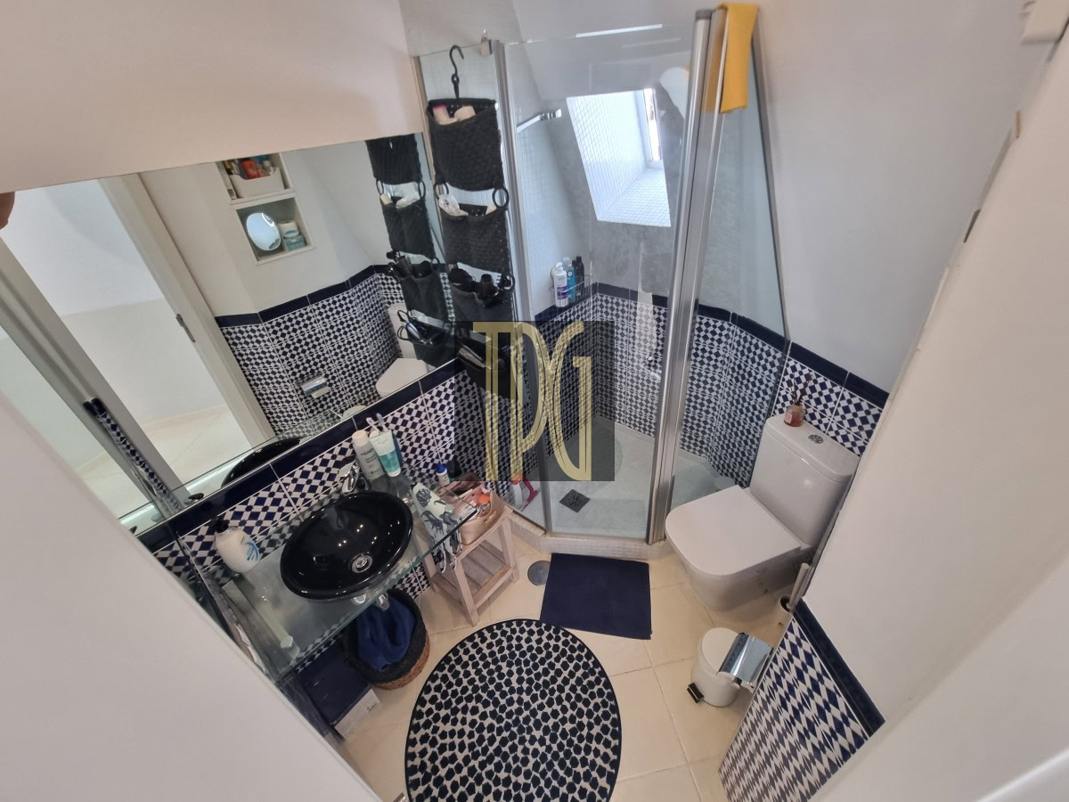 Small modern bathroom with a glass shower enclosure, black vessel sink on a glass counter, and a tiled blue‑white mosaic surround; round patterned rug nearby and a toilet against the wall.