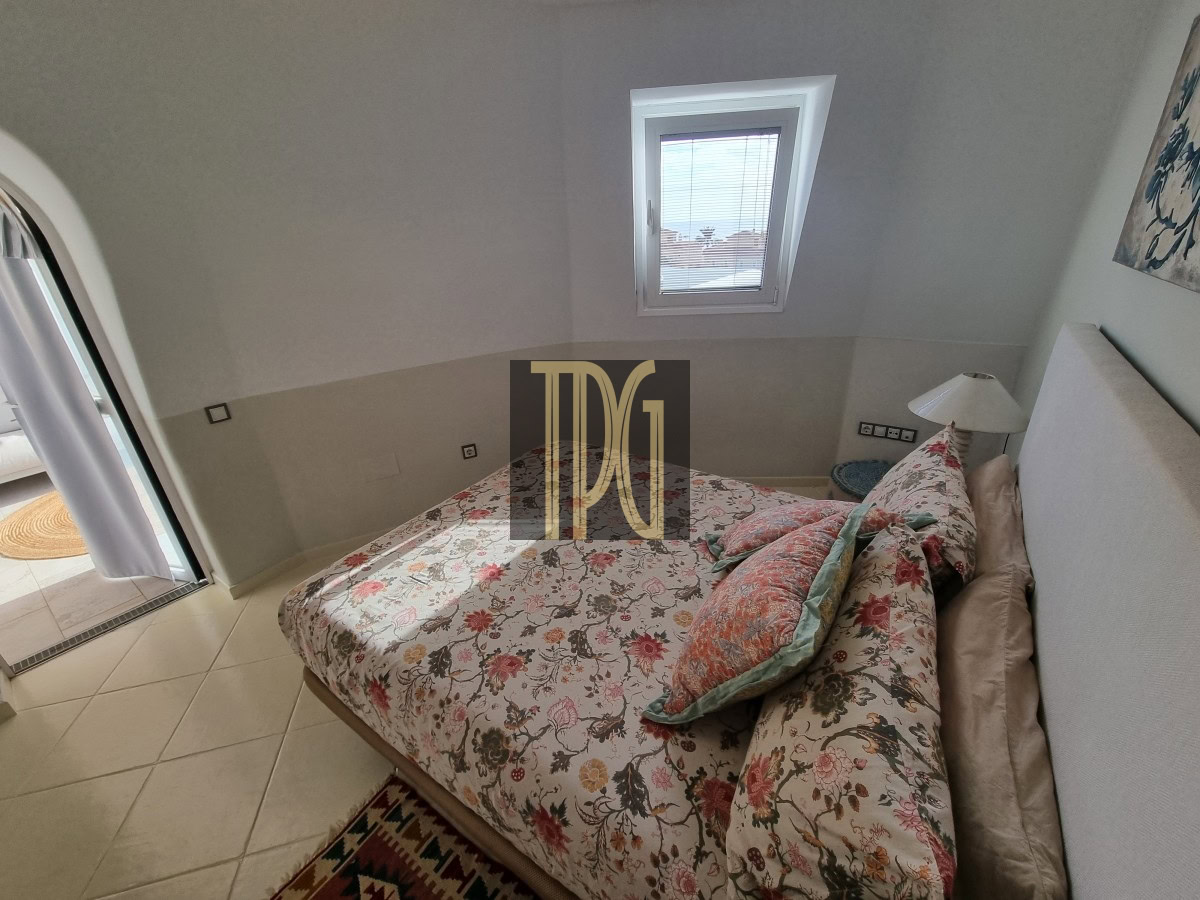 Small sunlit bedroom with a skylight above a floral bed, beige walls, and tiled floor; bouquet bedding and pillows create a cozy look.