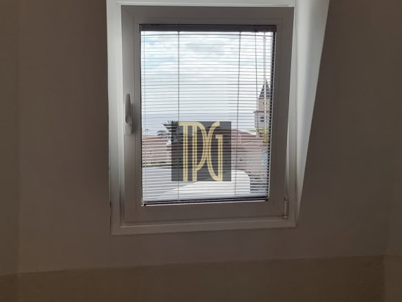 Small rectangular window with a white frame and horizontal blinds, showing rooftops and blue sky outside.
