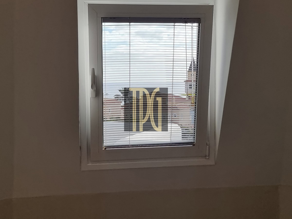 Small rectangular window with a white frame and horizontal blinds, showing rooftops and blue sky outside.