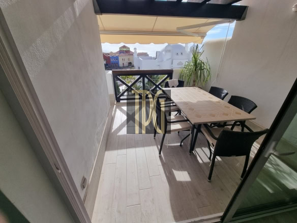 Outdoor balcony with beige tiled floor, rectangular table and six black wicker chairs, overlooking colorful buildings in the distance.