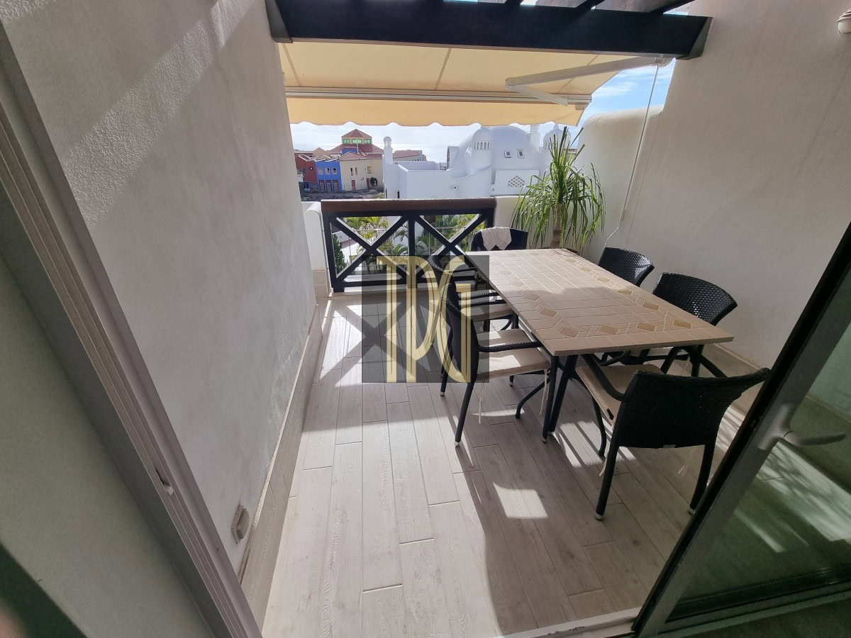 Outdoor balcony with beige tiled floor, rectangular table and six black wicker chairs, overlooking colorful buildings in the distance.