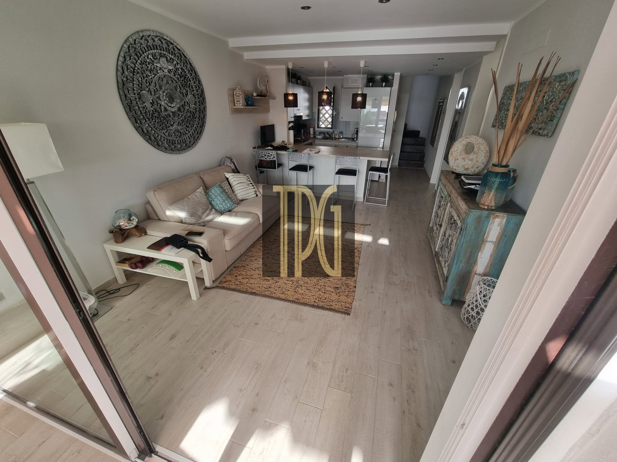 Open-plan living room with beige sofa and white side table, leading to a modern kitchen island and bar stools in the background.