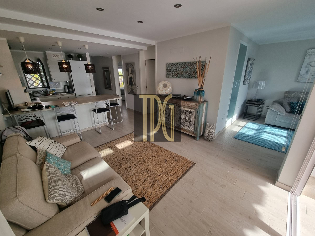 Open-plan living area with a beige sofa facing a front rug, view into a kitchen with an island and bar stools, and a glass door to a sunlit seating nook.