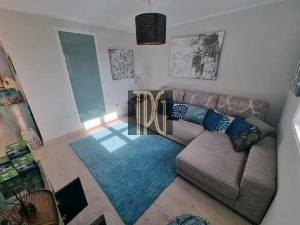 Cozy living room with a grey L-shaped sofa, blue cushions, and a blue rug under sunlight. Wall art and a black pendant light above.