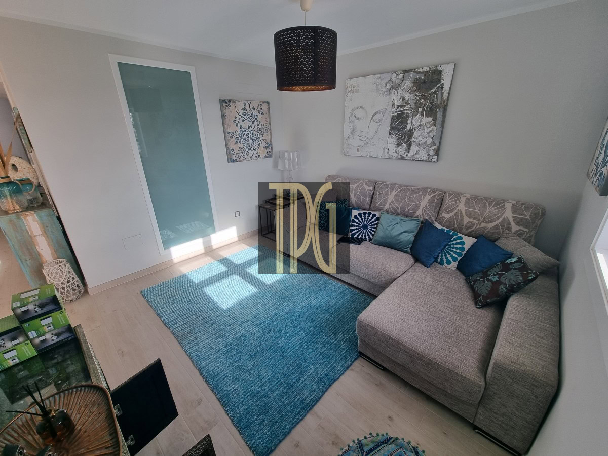 Cozy living room with a grey L-shaped sofa, blue cushions, and a blue rug under sunlight. Wall art and a black pendant light above.