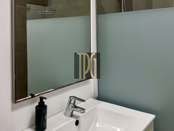 White bathroom sink with chrome faucet and a black soap bottle, mirror above, frosted glass shower panel to the right.