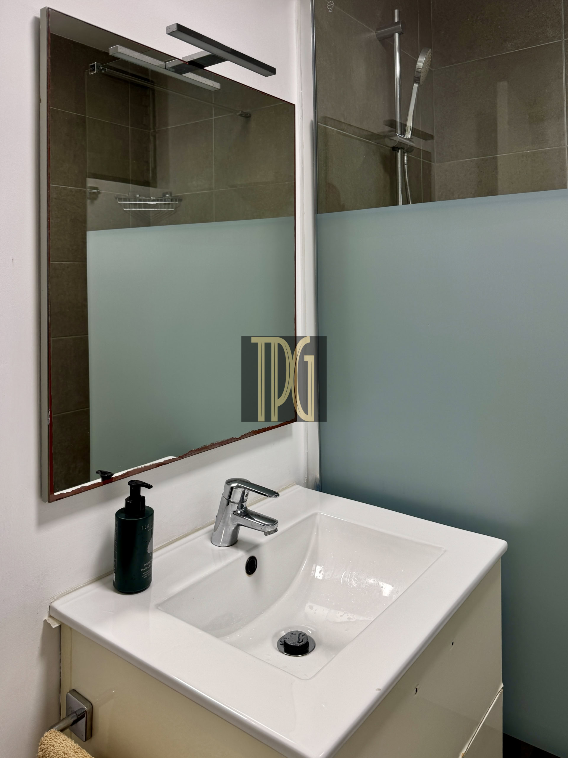 White bathroom sink with chrome faucet and a black soap bottle, mirror above, frosted glass shower panel to the right.