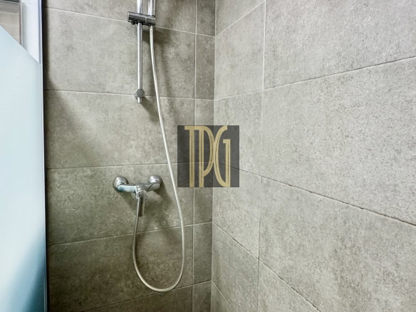 Shower with a chrome handheld showerhead on a vertical rail, mounted to gray tiled walls beside a glass panel.