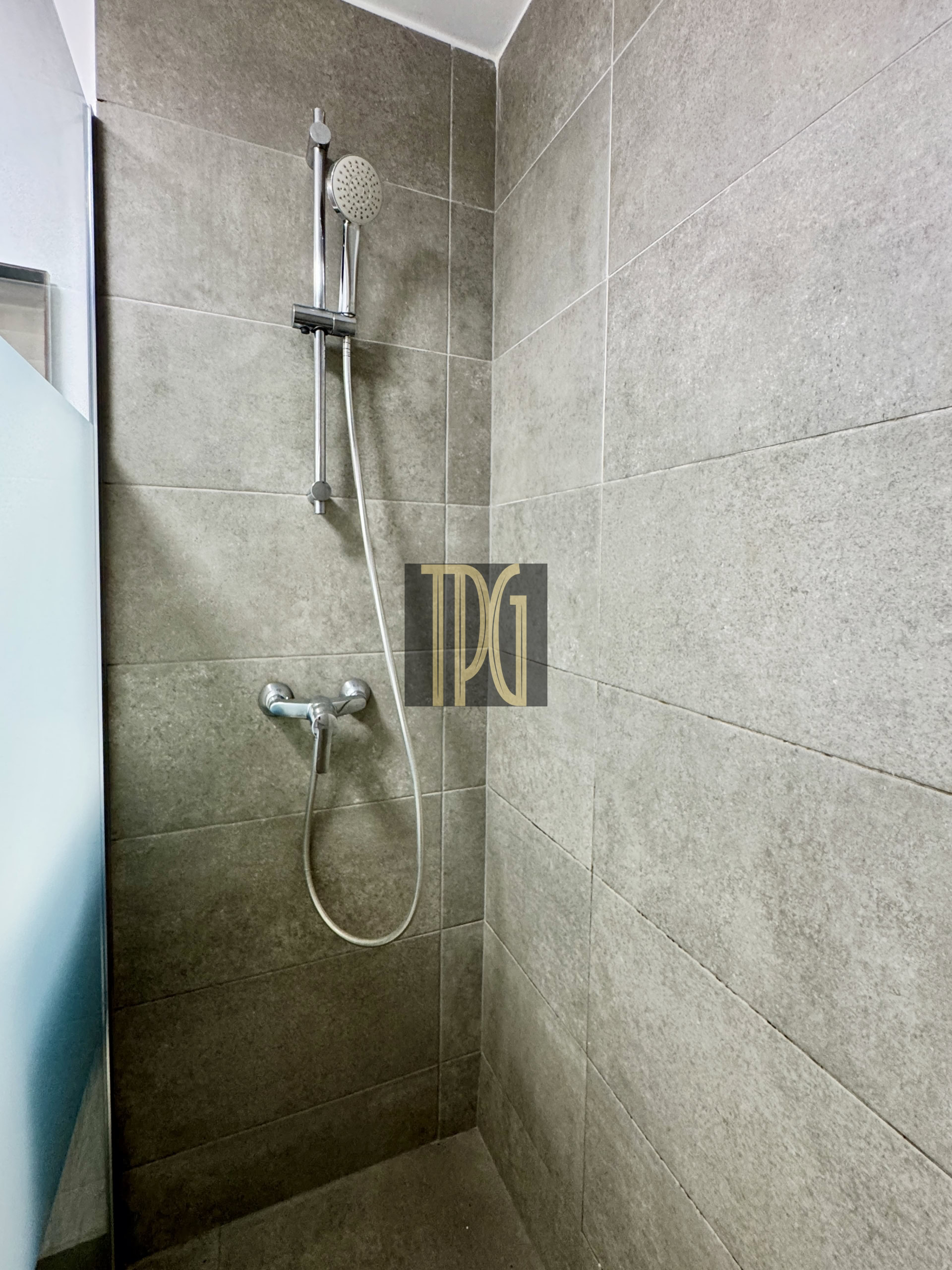 Shower with a chrome handheld showerhead on a vertical rail, mounted to gray tiled walls beside a glass panel.