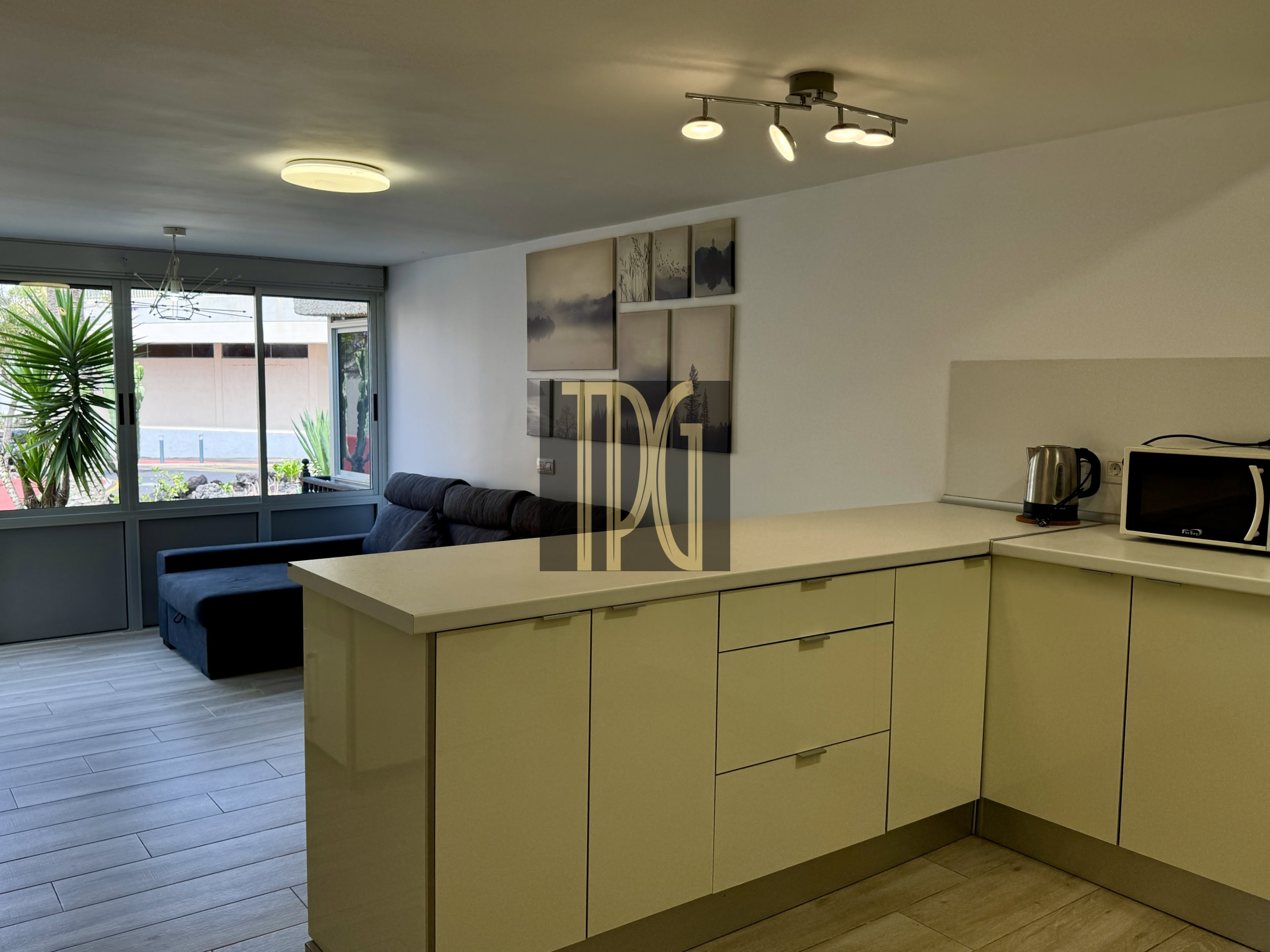 Open-plan living space with a beige kitchen island, glossy drawers, and a gray sofa under a wall of nature-themed multi-panel art; large windows and plants outside add greenery into the room.