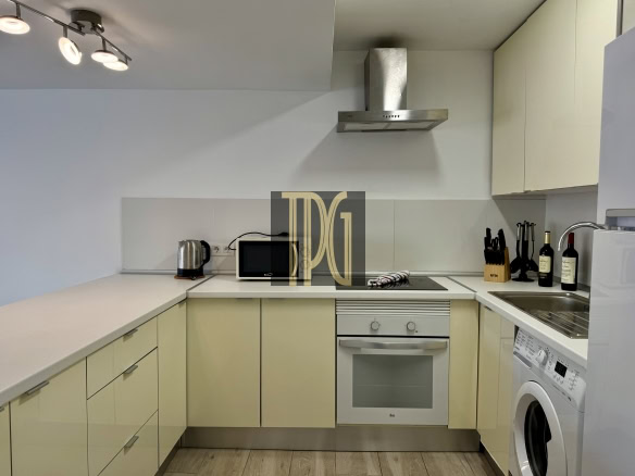 Modern L-shaped kitchen with pale cabinets, stainless steel range hood, built-in oven, microwave, and a washing machine visible on the right.
