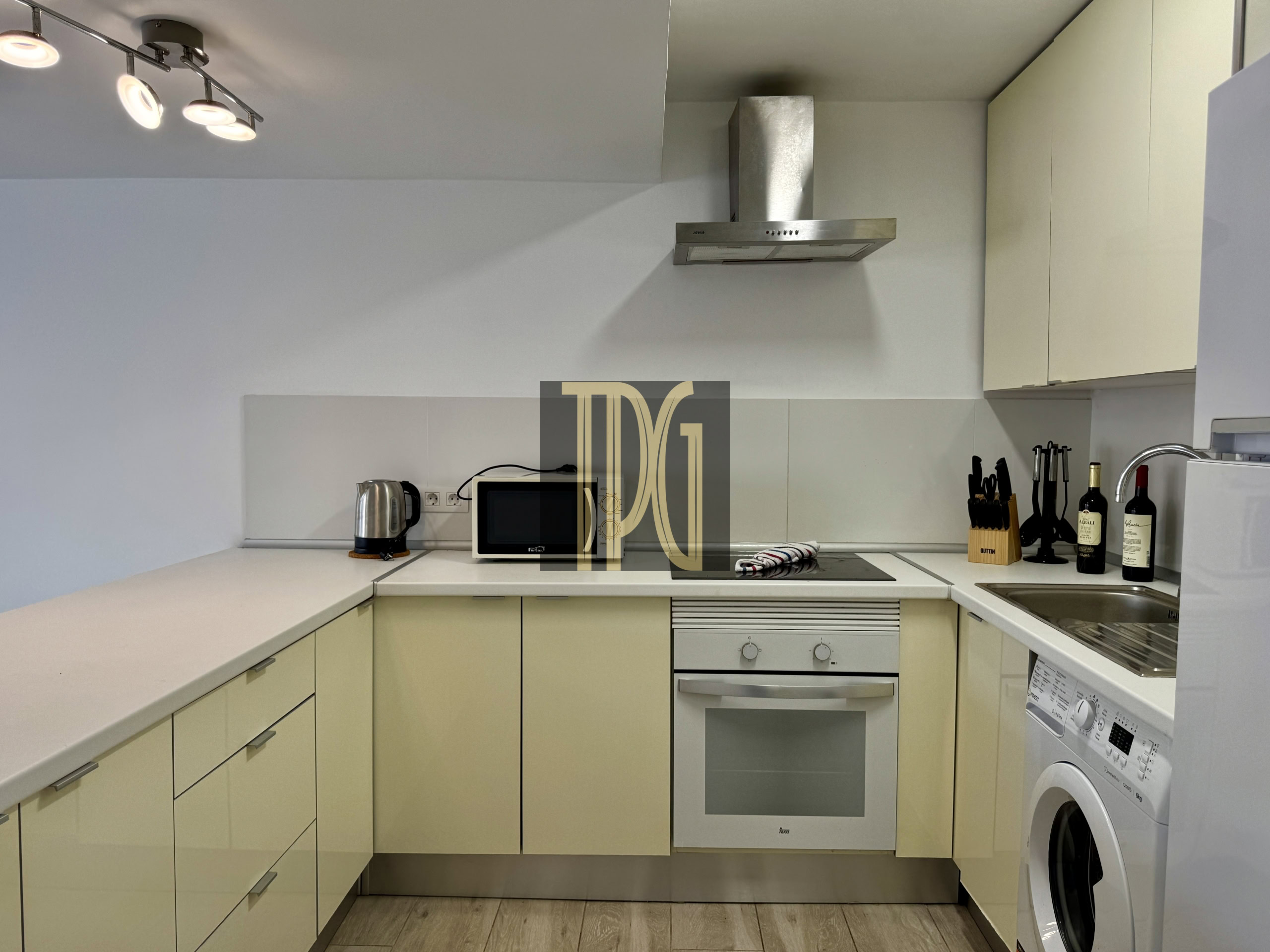 Modern L-shaped kitchen with pale cabinets, stainless steel range hood, built-in oven, microwave, and a washing machine visible on the right.