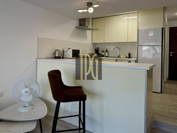 Modern kitchen with beige upper cabinets, stainless range hood, microwave, kettle, wine bottles, and a white refrigerator; a brown bar stool in front.