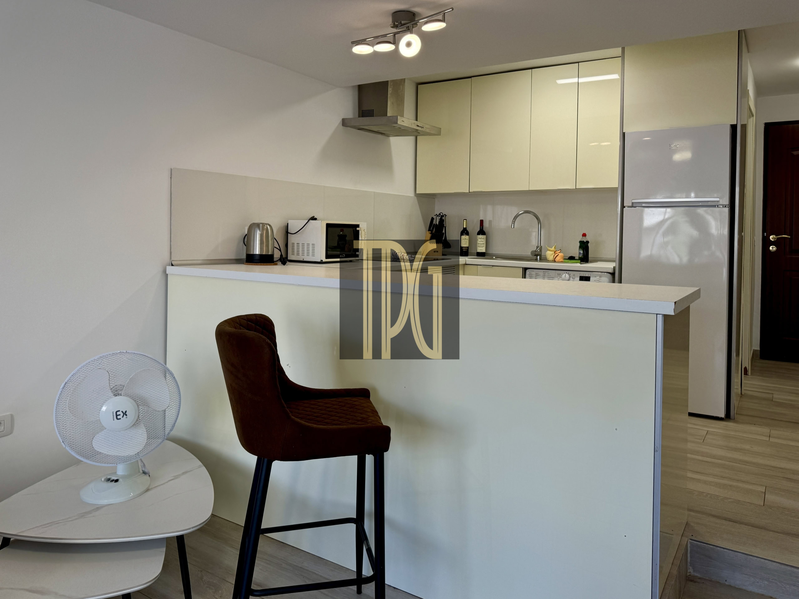Modern kitchen with beige upper cabinets, stainless range hood, microwave, kettle, wine bottles, and a white refrigerator; a brown bar stool in front.